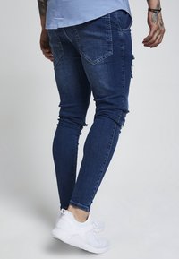 Man som bär mörkblå tajta slitna jeans och vita sneakers, stående mot en ljusgrå bakgrund.