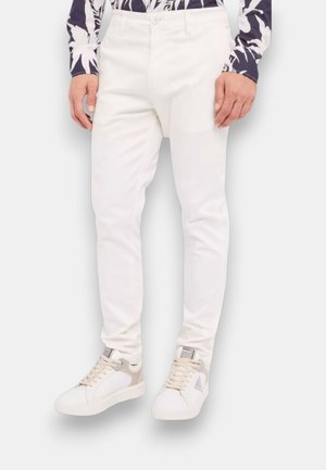 Uomo che indossa pantaloni bianchi slim fit e scarpe da ginnastica bianche con dettagli beige e grigi, in piedi davanti a uno sfondo semplice.