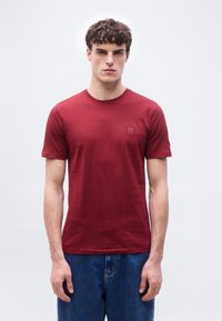 Νεαρός άνδρας που φορά ένα απλό κόκκινο κοντομάνικο μπλουζάκι με μικρό λογότυπο στο στήθος και μπλε τζιν, στέκεται μπροστά σε ανοιχτό γκρι φόντο.