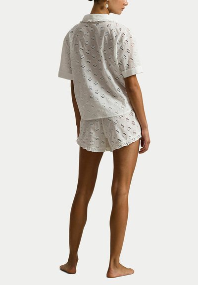 Vrouw draagt een witte, korte mouw eyelet-top en bijpassende korte broek, staat blootsvoets en lichtjes zijwaarts gedraaid tegen een egale achtergrond.