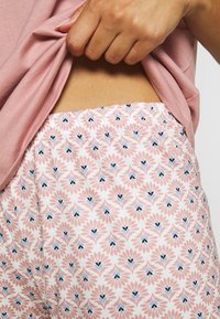 Lichtroze pyjamashorts met bloemenpatroon en elastische tailleband. Heeft herhalende blauw en zwarte accenten op een zachte, witte achtergrond.