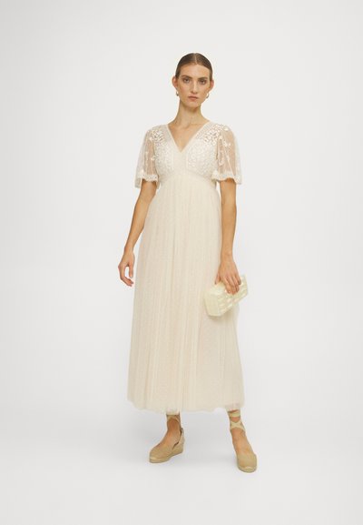 Abito maxi in crema chiaro con maniche a sbuffo sheer e corte, corpetto ricamato e gonna fluente e testurizzata, abbinato a una piccola pochette crema.