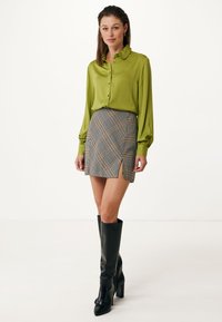 Camicia verde con bottoni e maniche lunghe, abbinata a una mini gonna a quadretti con spacco laterale, completata da stivali neri fino al ginocchio.