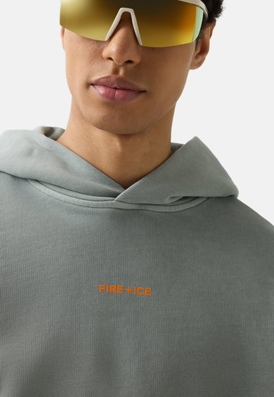 Junger Mann mit großen reflektierenden goldenen Sonnenbrillen und einem hellgrauen Hoodie mit orangefarbenem "FIRE+ICE" Schriftzug auf der Brust.