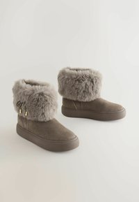 Bruine suède laarzen met een zachte, grijze nepbont manchet, voorzien van goudkleurige hardware-accenten en een platte rubberen zool voor stabiliteit.