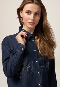 Camicia in denim scuro con chiusura a bottoni, caratterizzata da dettagli in paillettes argentate disposti in linee verticali e colletto classico. Texture liscia e maniche lunghe.