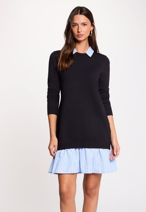 Kvinde iført sort langærmet sweaterkjole lagdelt over blå og hvide stribede skjortekjole med krave, stående mod en ensfarvet lys baggrund.