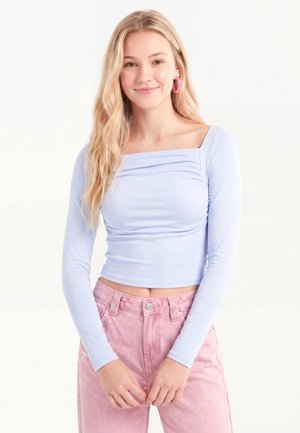 Ung kvinde med langt blondt hår iført en lyse lilla langærmet top og højttaljede lyserøde bukser, smilende med hænderne foldet sammen.