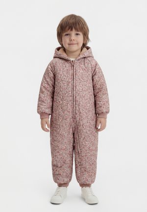 Noor laps seisab, seljas roosast mustriga kapuutsiga vooderdatud kombinesoon ja valged tossud, naeratab kergelt tavalise tausta ees.