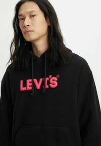 Felpa nera con logo "LEVI'S" rosso e rosa, cordini e tasca frontale. Texture morbida con vestibilità rilassata.
