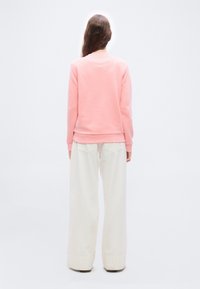 Άτομο με μακριά καστανά μαλλιά που φοράει ροζ φούτερ και φαρδιά λευκή παντελόνα, στέκεται με το πρόσωπο στραμμένο σε απλό γκρι ανοιχτό φόντο.