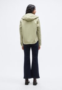 Ελαφρύ πράσινο-ελαιοχρώματος μπουφάν με κουκούλα, με ομαλή υφή και άνετη εφαρμογή, συνδυασμένο με σκούρα τζιν παντελόνια καμπάνα και καφέ μπότες με τακούνι.