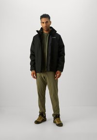 Jachetă impermeabilă neagră cu guler înalt, închidere cu fermoar pe față și logo. Cămașă și pantaloni de culoare verde măslinie, asortați cu pantofi de drumeție bej.