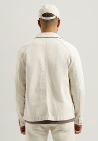 Lys beige denimjakke med klassisk krage, lange ermer og knappdetaljer, vist bakfra. Laget av bomull med en myk tekstur.