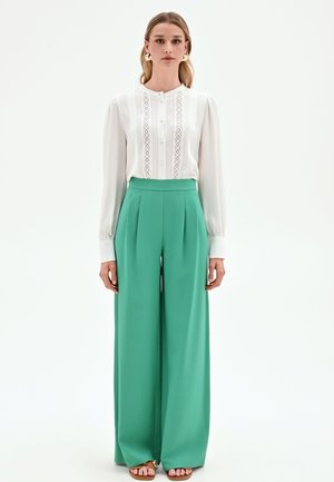 Donna in piedi, indossa una camicetta bianca a maniche lunghe con dettagli in pizzo e pantaloni verdi a vita alta e gamba larga, sandali marroni e orecchini dorati.