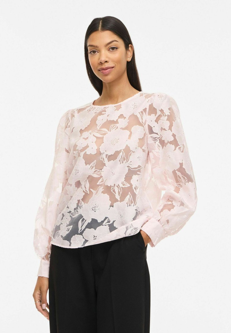 Femme portant un chemisier blanc transparent orné de broderies florales et de manches longues bouffantes, associée à un pantalon noir ajusté, posant avec une main dans la poche.