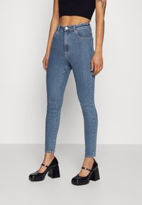 Jeans ajustados de tiro alto en azul con una textura de denim suave, que cuentan con un cierre frontal de botón y un diseño de cinco bolsillo, combinados con tacones negros de bloque.