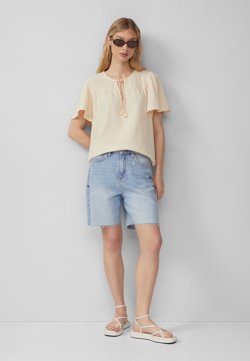 Top corto color beige chiaro a maniche corte con dettaglio a fiocco, abbinato a pantaloncini di jeans azzurri con orlo grezzo e sandali bianchi con cinturini.