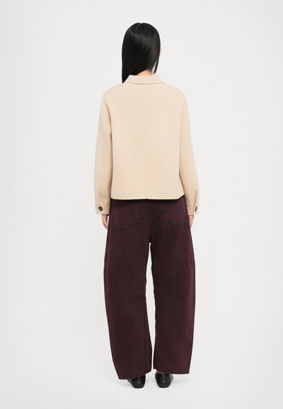 Chaqueta recortada beige con cuello, llevada sobre unos pantalones vaqueros anchos de pierna ancha en un oscuro burdeos. Los pantalones presentan detalles de costura visibles y bolsillos.