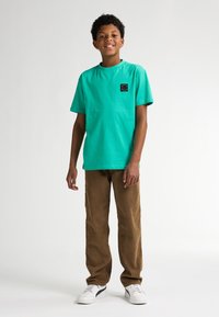 T-shirt di cotone turchese con maniche corte e un logo nero quadrato sul petto, abbinata a pantaloni di velluto a coste marroni e sneakers bianche.