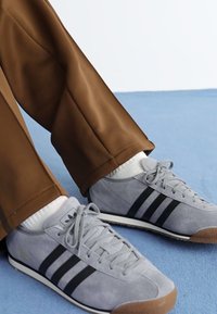 Pelēki zamšādi sporta apavi ar melnām svītrām, baltām zolem un gaiši pelēkām auklām; apvienoti ar brūniem biksēm un baltām zeķēm uz zilas virsmas.