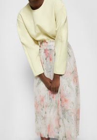 Femme portant un haut à manches longues jaune pâle et une jupe blanche transparente avec des motifs floraux roses et verts, se tenant devant un fond uni.