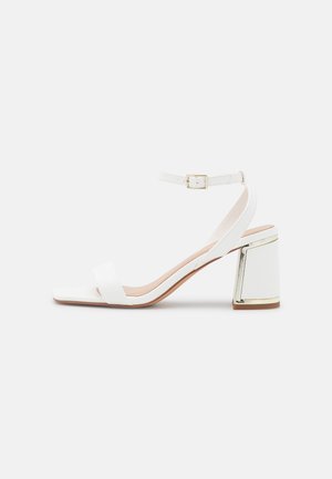 Sandale blanche à talons hauts avec un bout carré, une bride à la cheville et un talon bloc avec des accents dorés. Matériau en cuir lisse. Design minimal.