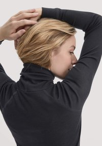 Schwarzer, figurbetonter Pullover mit Stehkragen, geripptem Design und geschwungenen Nähten. Das Haar des Models ist locker gestylt und zurückgehalten.