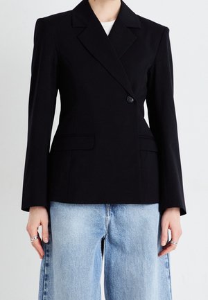 Czarna marynarka na jeden guzik z klapami z wycięciami, noszona na białej bluzce i jasnoniebieskich, szerokich, wysokich dżinsach, ręce opuszczone wzdłuż ciała z pierścieniami.