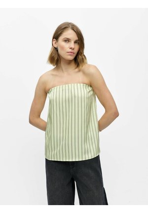 Woman wearing a strapless, pale green top with thin vertical stripes and dark wide-leg pants, standing with hands behind her back.