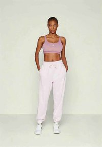 Mulher alta a usar um soutien desportivo mauve, calças de treino rosa claro e sapatilhas brancas, de pé com as mãos nos bolsos contra uma parede simples.