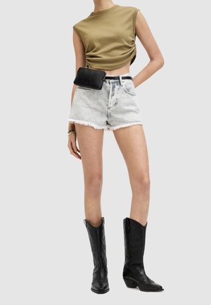 Frau trägt olivgrünes, gerafftes ärmelloses Crop-Top, hoch taillierte, hellblaue Denim-Shorts mit ausgefransten Kanten, schwarze Gürtel-Tasche und schwarze Cowboy-Stiefel.