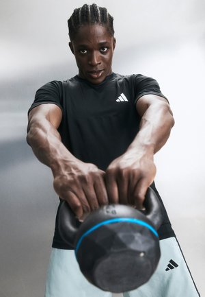 Személy két kézzel emel egy kettlebellt, fekete Adidas pólót és világos rövidnadrágot visel, és az erőnléti edzésre koncentrál.