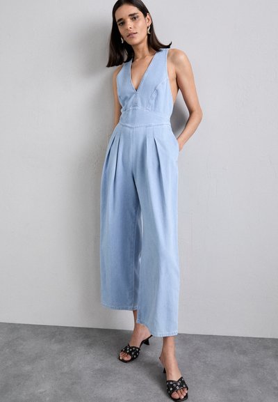 Combinaison en denim bleu clair à jambes larges, décolleté en V profond, poches latérales et détails plissés. Associée à des talons ouverts noirs avec des accents.