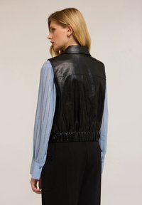 Czarna skórzana kamizelka bez rękawów z wysokim kołnierzem i elastycznym pasem, noszona na jasnoniebieskiej koszuli z dekoracyjnymi przeszyciami.
