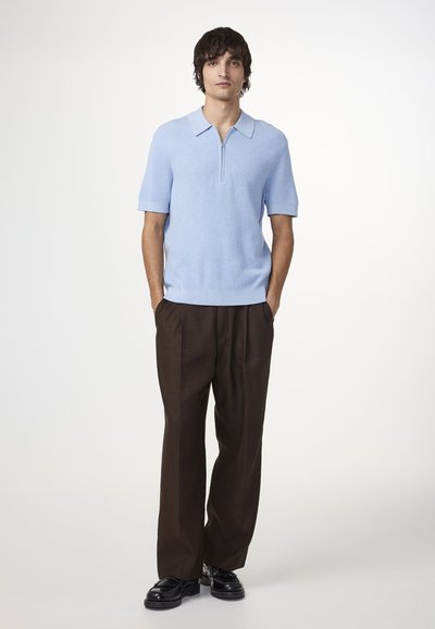Vaaleansininen lyhythihainen polo, jossa on vetoketjukaulus ja ribbitekstuuri, yhdistettynä rentoihin ruskeisiin housuihin ja mustiin kenkiin.