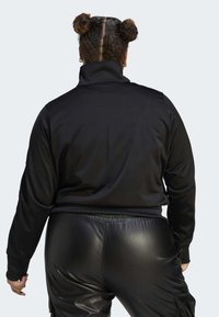 Osoba s dvěma drdoly, oblečená v černém vysokém límci a černých kožených kalhotách, zobrazená zezadu na jednobarevném pozadí.