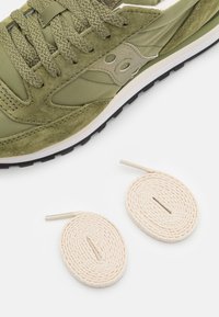 Olivgrüner Sneaker mit Obermaterial aus Wildleder und Nylon, der beige Akzente und flache olivgrüne Schnürsenkel aufweist. Inklusive zwei cremefarbenen ovalen Schnürsenkeln mit Metallspitzen.