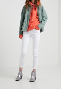 Jasně zelená denimová bunda, jasně oranžová mikina, bílé slim-fit džíny a metalické stříbrné kotníkové boty s černými akcenty na patách.