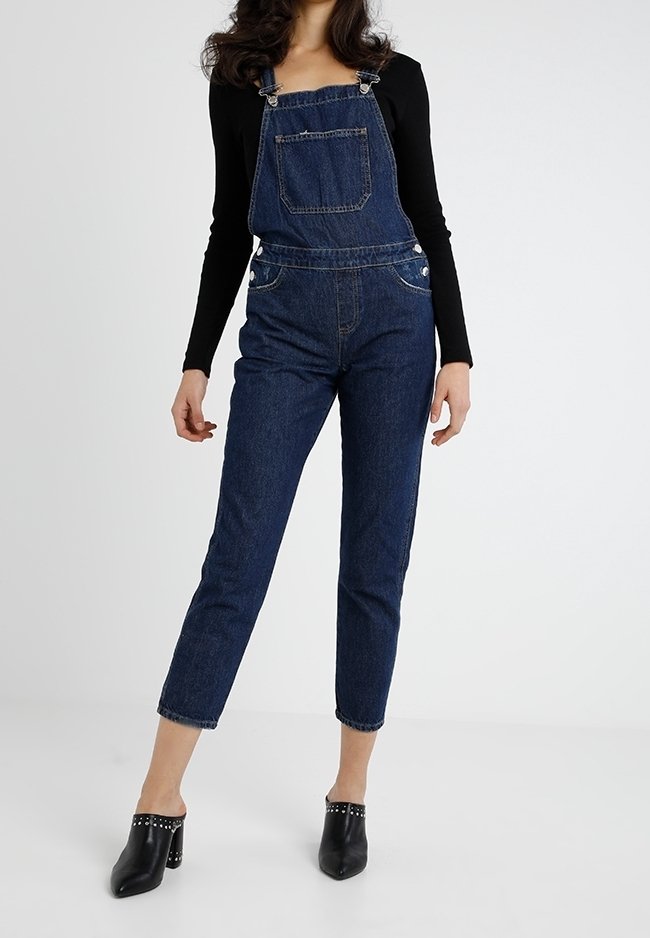 Femme portant une salopette en jean bleu foncé avec une poche devant, un haut noir à manches longues et des mules à talons noires cloutées, debout sur un sol blanc.