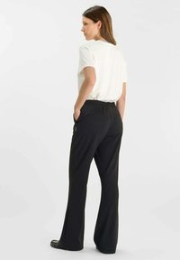 Vrouw met bruin haar, gekleed in een witte short-sleeve shirt dat in zwarte wijde broek is gestopt, staat met haar handen in haar zakken, met het gezicht naar links.