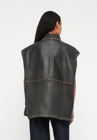 Persona con lunghi capelli ricci che indossa un gilet in pelle scuro senza maniche, visto da dietro contro uno sfondo bianco.