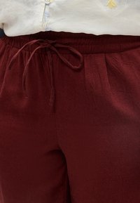 Κοντινή λήψη μαρόν παντελονιού με ελαστική μέση και δεμένο κορδόνι, φορεμένο με λευκό πουκάμισο με κουμπιά και κέντημα.