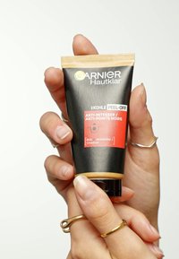 Tube en plastique noir avec un capuchon doré strié. Présentant une étiquette rouge avec du texte décrivant des propriétés anti-comédons. Tenue entre des doigts.