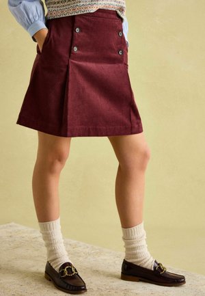 Maroon corduroy skirt with two side pockets and button accents. Worn with cream ribbed socks and dark loafers. Simple, structured design.