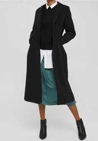 Persoon in een zwarte lange jas over een zwarte trui en een wit shirt, met een teal satijnen rok en zwarte enkellaarsjes, staand met de handen in de zakken.