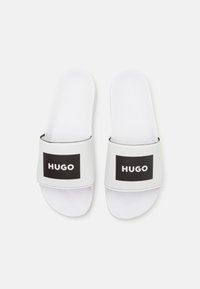 Hvite sklitt sandaler med en bred svart stropper som har ordet "HUGO" i fet hvit tekst, glatt tekstur og minimalistisk design.