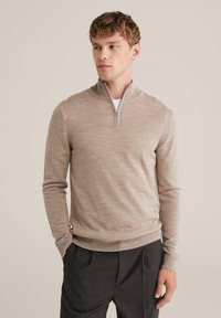 Beige zip-záras pulóver puha kötött anyagból, bordás gallérral és mandzsettákkal, sötét szürke nadrágokkal párosítva. Egyszerű és testre szabott dizájn.