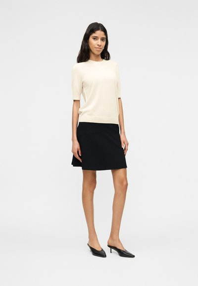 Femme debout portant un haut beige à manches courtes, une jupe noire jusqu'aux genoux et des chaussures noires à talons bas et bouts pointus, sur un fond uni.