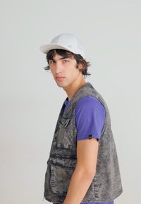 Vesta din denim gri, cu multiple buzunare, purtată peste un tricou mov. Modelul poartă o șapcă albă, cu fața ușor întoarsă spre lateral.